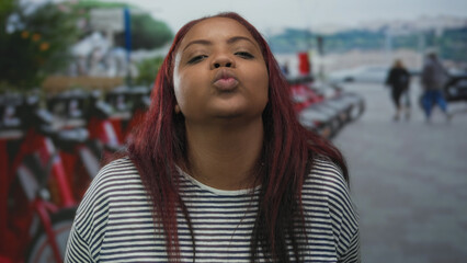 Young african american woman blowing a kiss at a row of red rental bicycles on busy urban street; affection.