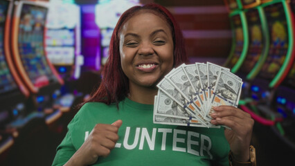 Woman holds a fan of hundred dollar bills with clenched fist at colorful casino slot machines indoors; victory.