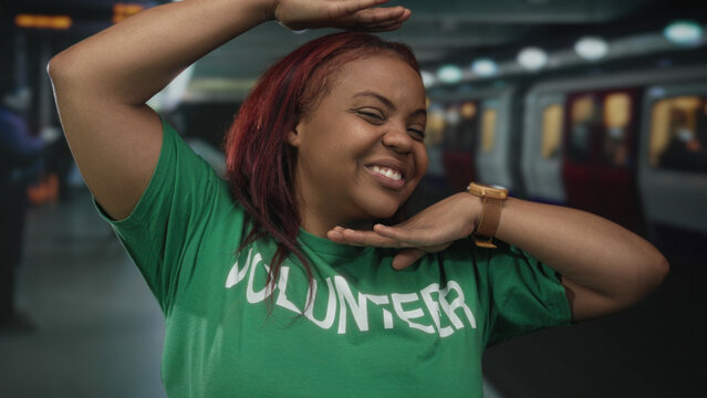 Volunteer in a green shirt smiles and frames her face with her hands beside a train in a building; playfulness community service volunteering.