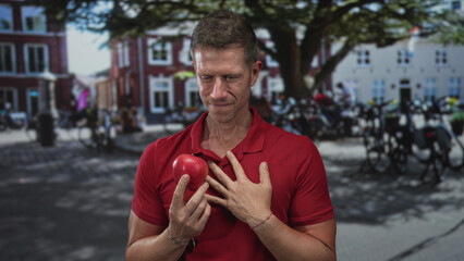 Man holding red apple, hand on chest, standing at house entrance on street in front of building, examining fruit with a pensive expression; quiet contemplation.