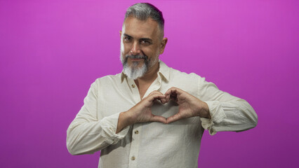 Obraz premium Middle-aged man with salt and pepper beard and rolled sleeves forms heart with hands at chest in a bright magenta pink studio, smiling and relaxed; affection.