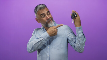 Man pointing finger to car keys while holding key fob and smiling, standing in purple studio; confidence car sale.