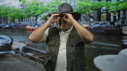 Man in fishing vest and hat using hands as binoculars peering toward canal boats on a street;...