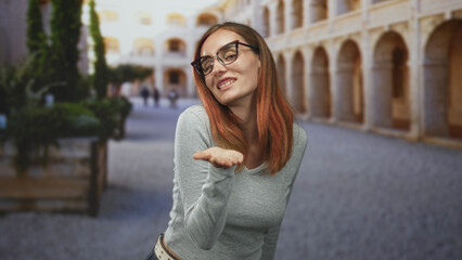 Obraz premium Woman redhead wearing a sweater with glasses blowing a kiss, palm extended in building courtyard; playful affection.