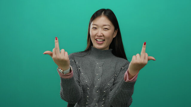 Woman smiling in a gray sweater gestures with middle fingers against a vibrant green background, capturing a bold expression and confident demeanor, showcasing casual attire.