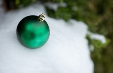 Green shiny Christmas ball ornament resting on fresh snow with natural blurred evergreen background. Minimal winter holiday decoration with copy space, suitable for Christmas design and marketing.