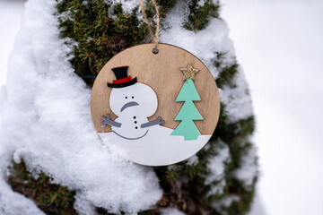 Wooden snowman ornament hanging on evergreen tree covered with snow. Natural outdoor winter decoration with rustic style, suitable for seasonal design, winter backgrounds, holidays and greeting cards.