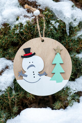 Wooden snowman ornament hanging on evergreen tree with snow and icicles. Natural winter decoration outdoors, showing melting snow, suitable for seasonal design, greeting cards and winter backgrounds.