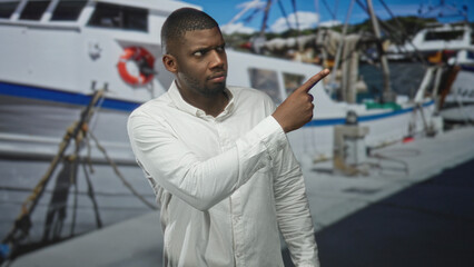 Man points finger at boat on dock in studio, wearing white shirt and beard, turns head with steady gaze; curiosity exploration.