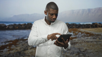 Man tapping tablet with right hand, holding device with left and making a raised hand gesture in studio with seaside rock shore backdrop; concentration.