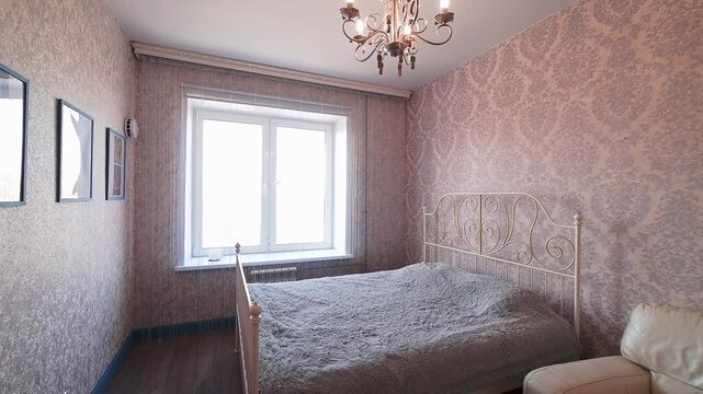 cozy bedroom with pink patterned wallpaper, a white metal bed frame, a chandelier, a large window with curtains, framed pictures on the wall, and a sofa corner