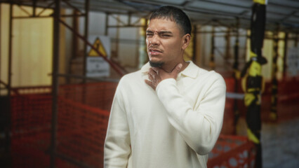 Young hispanic man clutching throat with hand, wincing amid construction scaffolding inside a...
