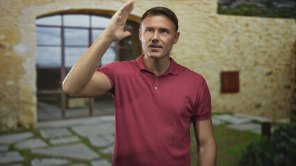 Fototapeta premium Handsome hispanic man touching forehead with palm and closed eyes, wearing red polo standing before an old stone building entrance; relief.