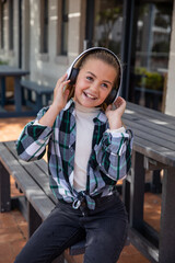 Girl child sitting on wooden picnic bench in courtyard with wireless headphones and smiling