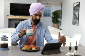 Mid adult indian man wearing turban working on tablet at kitchen island with coffee, toast