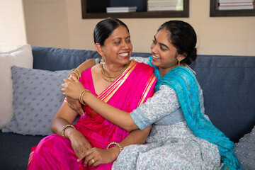 Indian mother in sari and daughter sharing embrace and wearing gold jewelry on couch in living room