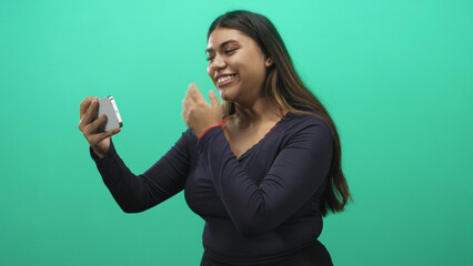Woman holds smartphone at arm's length, smiles and touches mouth while gesturing with other hand against green screen studio; joy connection.