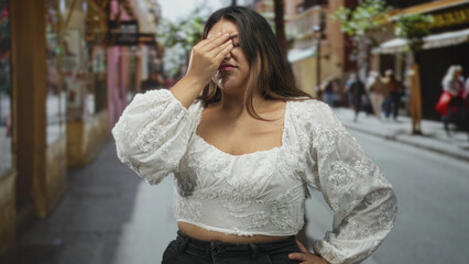 Woman with hand on forehead for headache on street wearing white embroidered blouse, wincing and rubbing temples; stress fatigue frustration.