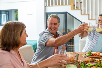 Senior friends toasting with glasses at home dining table with glass pitcher salad, vegetables