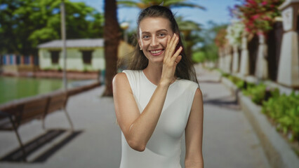 Fototapeta premium Woman with hand to cheek smiling and laughing on a sunny street by a green park walkway with bench and palm tree; happiness playfulness.