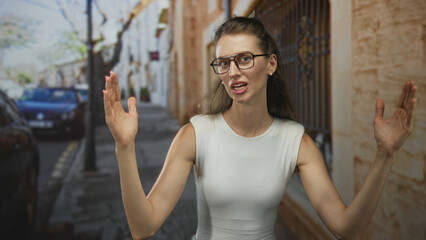 Obraz premium Woman on a sunlit street with hands raised and expressive face, wearing glasses and sleeveless white top; surprise.