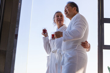 Senior couple standing by glass window in spa wearing bathrobes holding glass mugs with tea