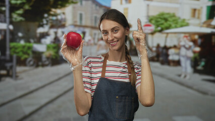 Woman holding red apple with gloved hands wearing apron and striped shirt points finger to apple in busy street; healthy confidence.