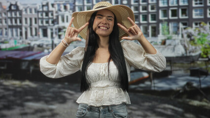 Fototapeta premium Young woman smiling and making rock hand gesture with fingers, wearing straw hat and white blouse with bare hands visible on street beside canal buildings; playful.