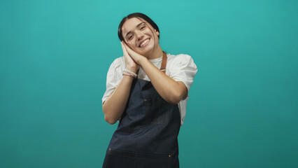 Woman in denim apron presses hands together at cheek in a studio, smiling with closed eyes; playful joy.
