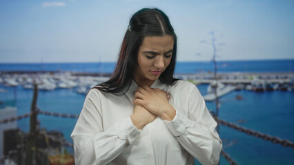 Fototapeta premium Woman with hands on chest standing at a port with boats in the background, wearing a white shirt, demonstrating an emotional and reflective moment outdoors by the sea.
