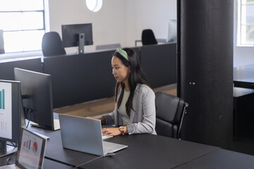 Chinese asian woman wearing grey blazer working on laptop at office with chart monitor