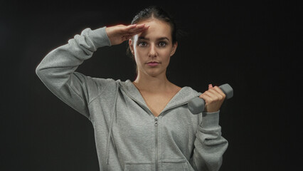 Young hispanic woman holding gray dumbbell and saluting with hand to forehead in studio against...