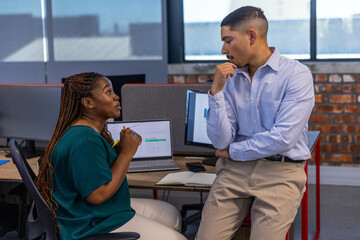 Diverse coworkers discussing data charts on laptop in office with monitors, notebook, smartphone