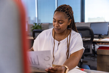 African american woman sitting at desk in office reviewing printed charts