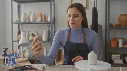 Woman holds ceramic vase and smartphone, hand visible while inspecting pottery in studio; creativity focus.