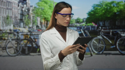 Woman scientist in white lab coat and safety goggles holding tablet on street in amsterdam by canal and bicycles; urban research focus.