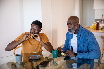 Senior african american father and son sitting at kitchen table eating sandwich, smartphone