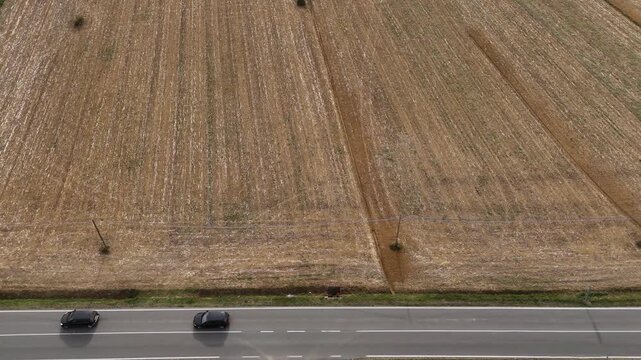 High-angle winter aerial of a rural Italian highway. Ploughed earth-toned fields with rhythmic vertical patterns parallel a paved road as dark vehicles travel through the agricultural heartland.