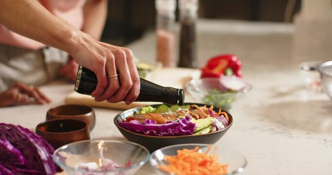 Hand is touching kitchen counter then lifting dark bottle and pouring dressing over salad to coat