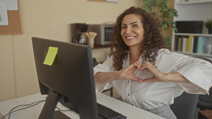 Hispanic woman forms heart shape with hands in front of computer monitor at office desk; love...