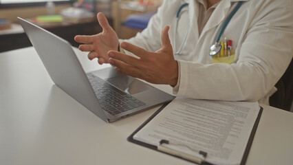 Doctor man in white coat gestures open hands over laptop and clipboard document at indoor clinic...