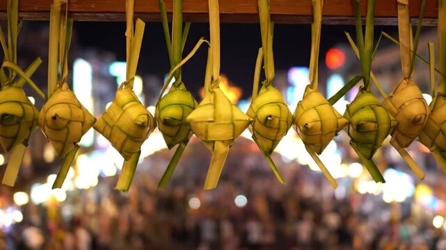 Close-up of traditional ketupat hanging in a row at a bustling night market with blurred crowd background.