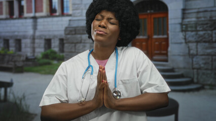 African american woman doctor wearing white scrubs and stethoscope pressing palms together at...