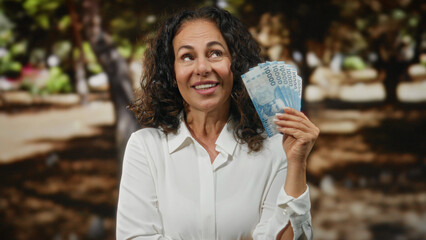Woman smiling while holding chilean pesos in a park, showcasing currency with a joyful expression outdoors.