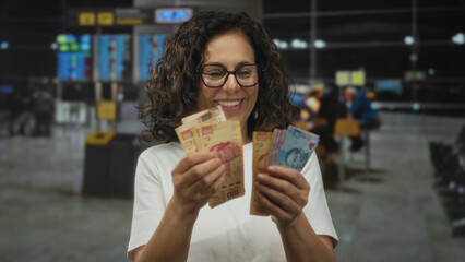 Woman smiling counting mexican pesos indoors at airport terminal, conveying travel finance concept with happiness and anticipation.