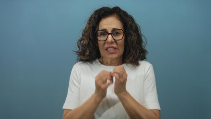 Middle aged hispanic woman anxiously bites her fingernails in front of isolated blue studio wall;...
