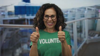Smiling hispanic woman in green volunteer shirt giving thumbs up on a seaside boat outdoors with blurred background emphasizing positivity.