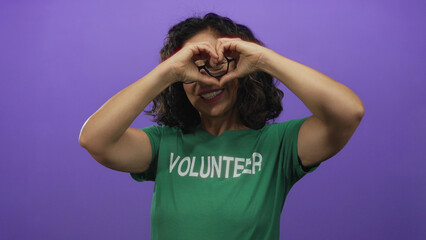 Woman in green uniform with glasses making heart symbol stands in front of purple wall highlighting community support and volunteerism.