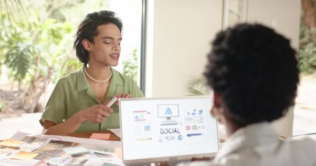 Diverse coworkers exchanging sticky notes at desk refining social graphics displaying on laptop