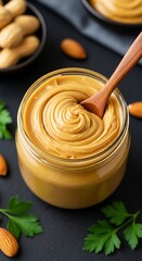 Creamy peanut butter in glass jar with wooden spoon surrounded by almonds and parsley on dark background